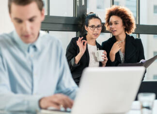 Ali opravljanje na delovnem mestu dejansko koristi? Znanost pravi … Two young women laughing while talking in the office behind their male colleague