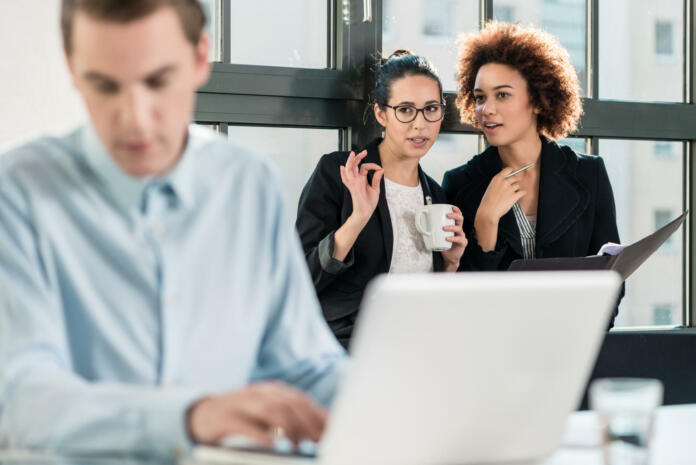 Two young women laughing while talking in the office behind their male colleague