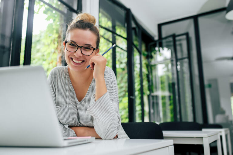 Portrait of smiling young female student in eyeglasses sitting in modern library.