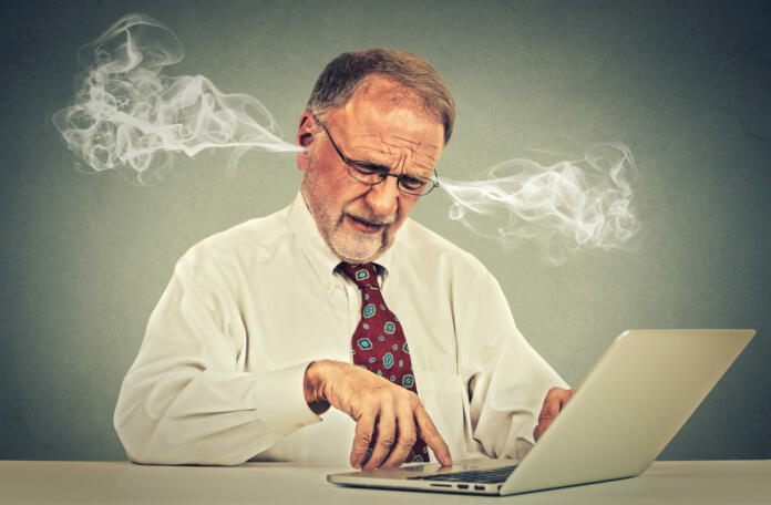 Stressed elderly old man using computer blowing steam from ears. Frustrated guy sitting at table working on laptop isolated on gray wall background. Senior people and technology concept