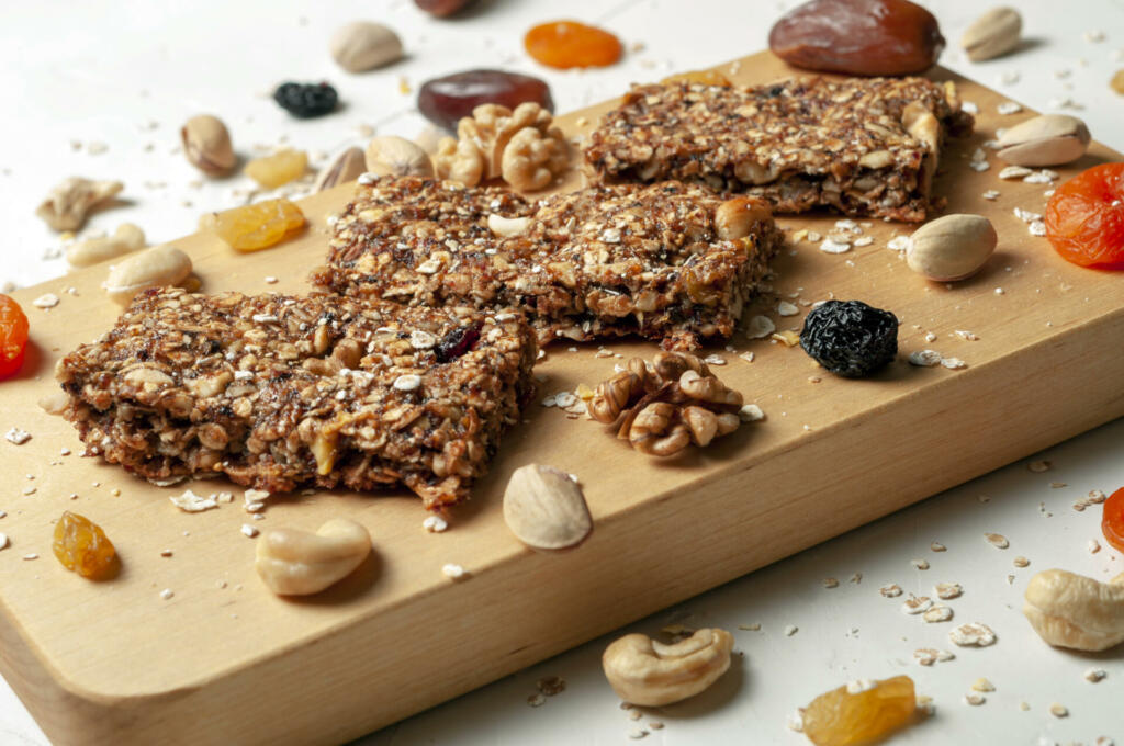 bar with granola, dried fruits and nuts on a light wooden board with dried fruits, nuts and cereals scattered nearby, close-up.