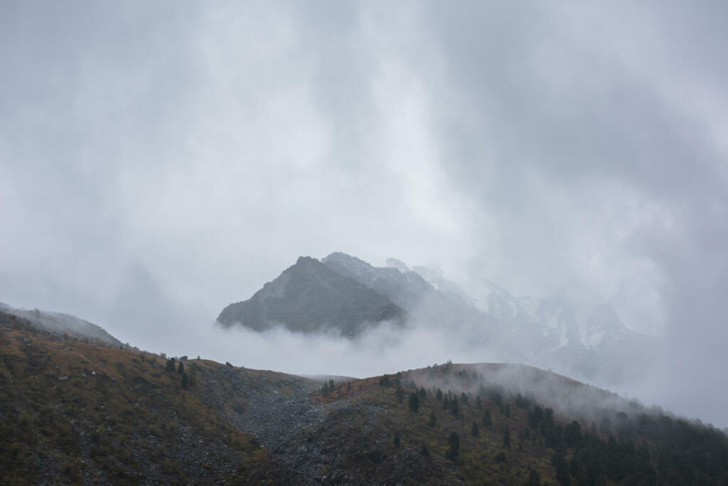 Dark atmospheric landscape with high mountain silhouettes in dense fog in rainy weather. Snowy rocky mountain top above hills in thick fog in dramatic overcast. Black rocks in low clouds during rain.