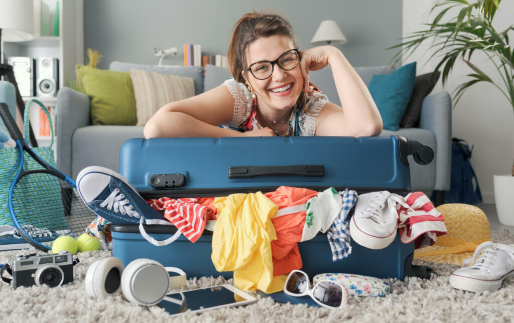 Happy young woman packing for a trip, she is leaning on her full trolley suitcase and smiling