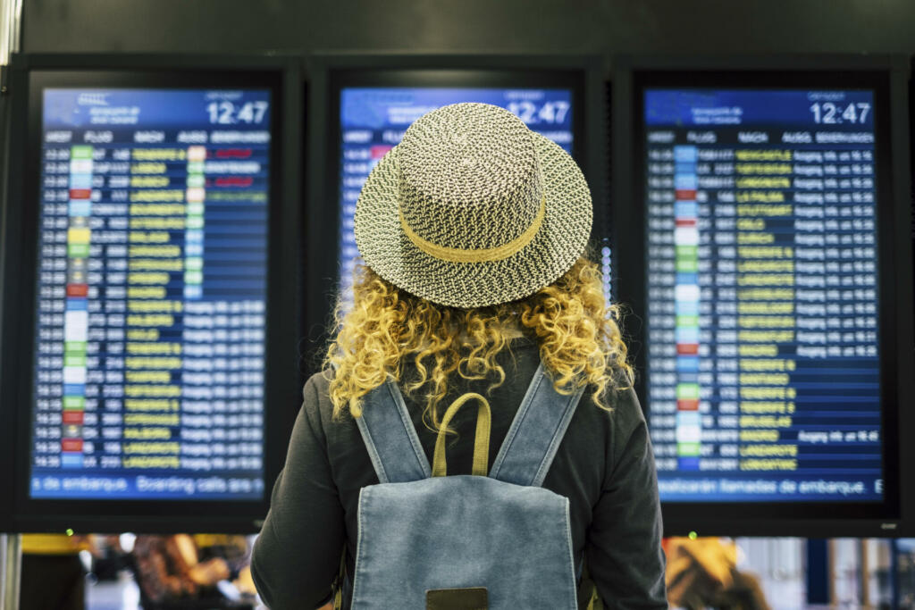 Travel people in airport or train station concept - view from rear of a blonde curly female woman with blue backpack looking and checking time departures or arrivals on the displays screen