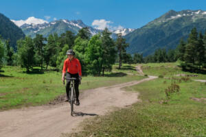A young smiling girl on a cyclocross bike rides along a winding mountain road against a background of green forest and mountains with glaciers and snow on the tops