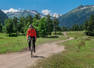 Je to najlepši kolesarski dogodek v Sloveniji? A young smiling girl on a cyclocross bike rides along a winding mountain road against a background of green forest and mountains with glaciers and snow on the tops