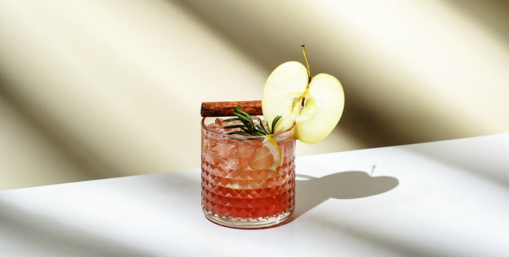Apple pie alcoholic cocktail drink with vodka, apple liqueur, cinnamon, lemon, garnished with rosemary. Light beige background, hard light, shadow pattern, banner