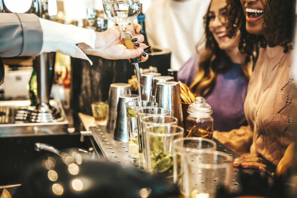 Bartender pouring alcohol from the bottle into the glasses - Happy friends group hanging out on weekend night at cocktail bar venue - Life style concept with barman making drinks and serves customers