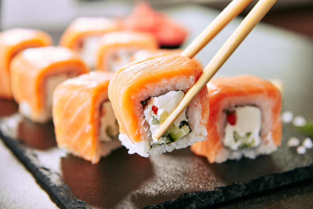 Chopstick holding Sushi Rolls Set with Salmon and Cream Cheese and Cuccumber on Black Slate Plate Close Up. Uramaki, Nori Maki or Futomaki Sushi with Trout Fillets, Soy Sauce and Wasabi