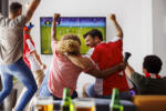 Group of young friends having fun watching football match on TV, drinking beer and cheering; football fans watching game at home celebrating after their team scoring a goal