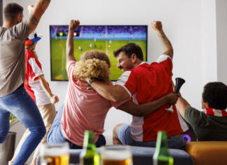 Tako gledanje športa vpliva na nas in naše življenje Group of young friends having fun watching football match on TV, drinking beer and cheering; football fans watching game at home celebrating after their team scoring a goal