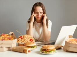 Indoor shot of sick unhealthy woman sitting in front of laptop among junk food, having terrible headache, being tire and exhausted, isolated over gray background.