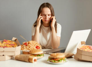 Tako si z zanič hrano uničujemo spomin (Raziskava) Indoor shot of sick unhealthy woman sitting in front of laptop among junk food, having terrible headache, being tire and exhausted, isolated over gray background.