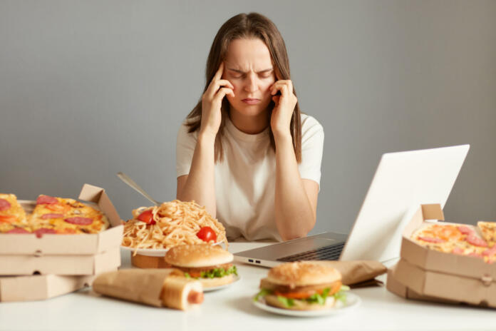 Indoor shot of sick unhealthy woman sitting in front of laptop among junk food, having terrible headache, being tire and exhausted, isolated over gray background.