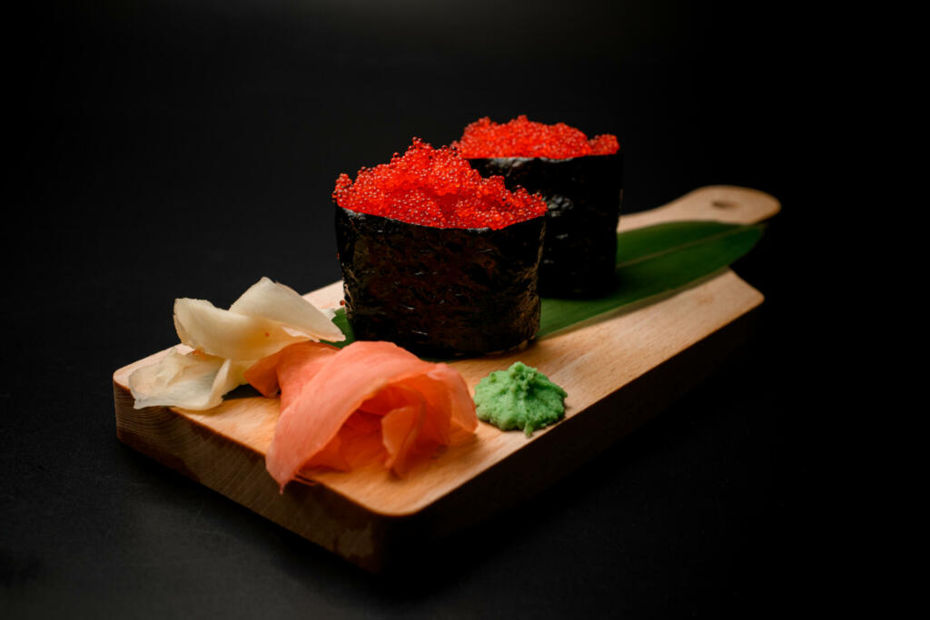 Luminous bright red tobiko delicately arranged on a leaf, accompanied by zesty ginger and pungent wasabi for a delightful flavor trio.
