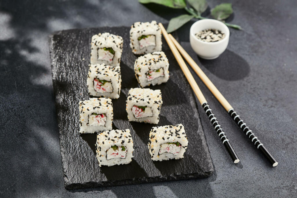 Maki sushi on dark slate. California maki with sesame. Sushi roll with crab, cucumber inside, sesame outside. Style concept japanese menu with black background, leaves and hard shadow