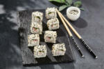 Maki sushi on dark slate. California maki with sesame. Sushi roll with crab, cucumber inside, sesame outside. Style concept japanese menu with black background, leaves and hard shadow