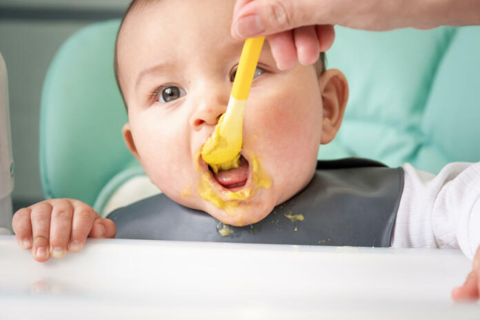 Mom feeds the baby with a spoon of vegetable puree at the children's feeding table. Baby's appetite, healthy nutrition, introduction of complementary foods. Copyspace, mock up