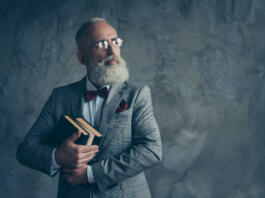 Portrait with copy space of successful, attractive, serious millionaire in jacket, glasses, holding three books in arms, with serious expression looking to the side over gray background