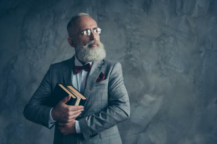 Portrait with copy space of successful, attractive, serious millionaire in jacket, glasses, holding three books in arms, with serious expression looking to the side over gray background