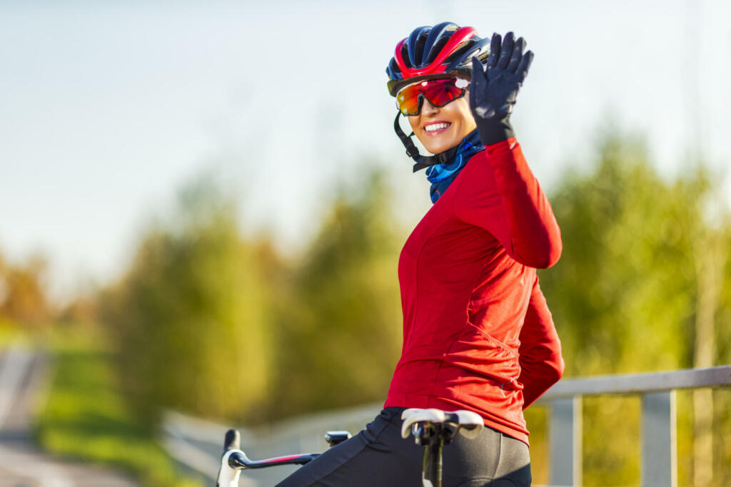 Winsome Caucasian Female Cyclist In Sport Outfit Posing On Road Bike With Lifted Hand While Waiving Outdoors Over Autumn Background. Horizontal Composition