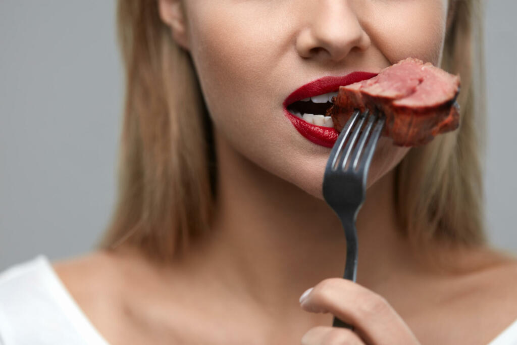 Woman Eating Meat. Closeup of Healthy Hungry Girl With Beautiful Face, Red Lips Eats Delicious Grilled Meat. Female Mouth Biting Piece Of Tasty Beef Steak On Fork. Nutrition Concept. High Resolution