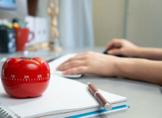 Najboljša tehnika učenja in dela, ki žanje uspehe na vseh področjih A selective focus shot of a kitchen timer in the form of a tomato on a notebook with a female working on her computer in the background
