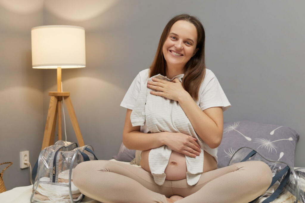 Adorable regnant woman preparing baby clothes for the maternity hospital embracing little kid's attire getting the nursery ready for the new arrival in home interior