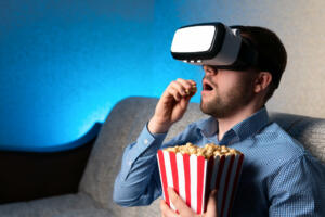Adult male in VR goggles eating fresh popcorn while sitting on comfortable sofa in stylish room