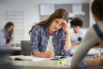 Anxious young woman with hand on head feeling tired while studying at school in classroom. College student suffering from headache in library preparing for test. Portrait of troubled and stressed girl doing exam that doesn"u2019t know the answers.