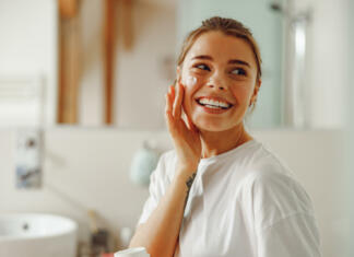 3 načini večerne nege obraza, ki so nujni za mladostniški videz Happy woman applying hydrating moisturizer on her face sitting in bathroom. Home beauty routine