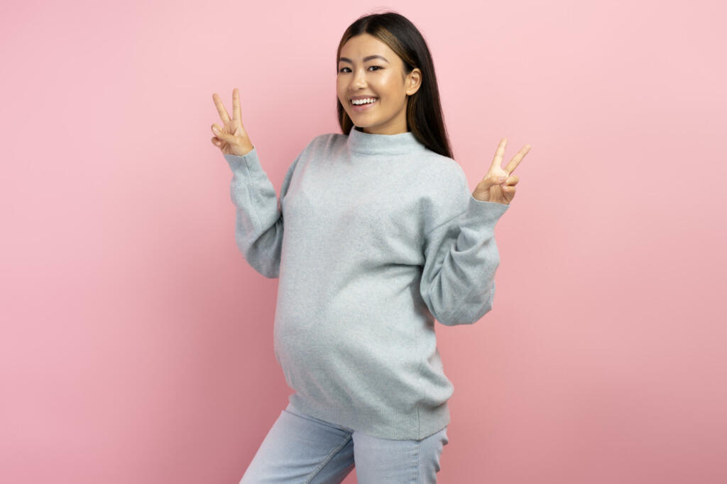 Pregnant woman wearing winter sweater drinking glass of water isolated on pink background. Water balance during pregnancy concept