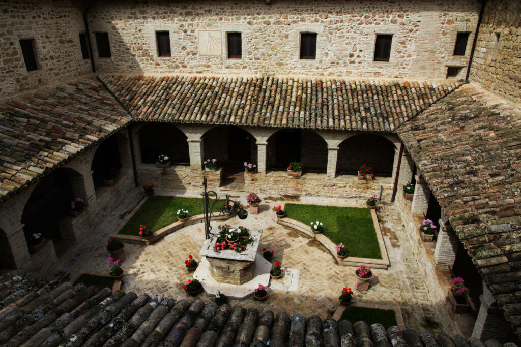 The courtyard of San Damiano, where St. Clare (Santa Clara) of Assisi lived out her days. From the window in this cloister, St. Clare had her last viewing of St. Francis of Assisi. The courtyard is empty. There is a sundial on the wall in the upper middle. Flowers decorate the courtyard, there are roman style shingles.