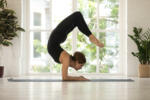 Woman in black sportswear performing Vrischikasana or Scorpion yoga asana near window green trees summer sunny morning view improve blood flow to brain stretches back develops sense of balance concept.