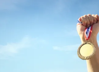 Kdo ima največ medalj na olimpijskih igrah? Gold medal winner held in athlete hand raised against blue sky background concept for winning, success and victory