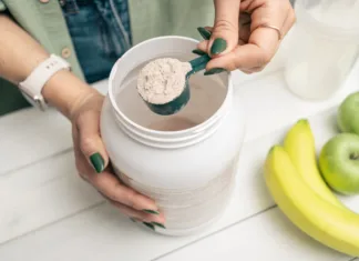 Najpogostejši miti o proteinih: Držijo ali ne? Woman in jeans and shirt holding measuring spoon with portion whey protein powder above plastic jar on white wooden table with shaker, banana and apple fruit. Process of making protein drink.