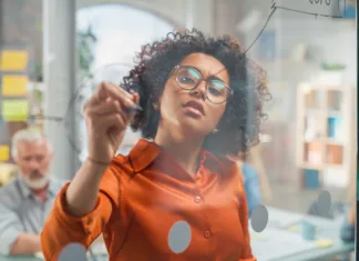 Kako biti kreativen na delovnem mestu? Portrait of Black Female Manager Writing on Glass Board and Brainstorming with Team in a Stylish Meeting Room in Creative Office. Team of Multi-Ethnic Professionals Focused on a Presentation