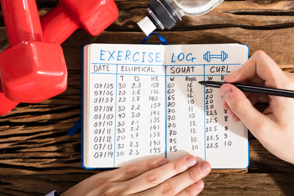 Elevated View Of A Hand Filling Exercise Log On Notebook With Exercise Equipment On Desk