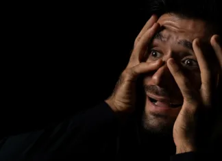 Zakaj tako radi gledamo grozljivke? Frightened young beard man afraid scared and terrified with fear expression face He put hands on face Young guy were shock and panic in the horror dark room Emotional and facial expression concept