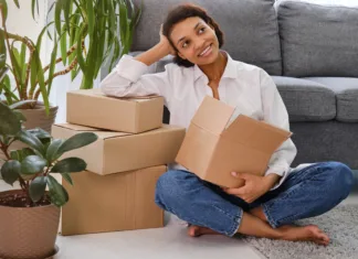 Kako ustvariti udobno in funkcionalno študentsko stanovanje: Nasveti za notranjo ureditev Happy young woman sitting in new apartment and raising arms in joy after moving in. Joyful and excited african girl moving to new home. Black woman sitting on floor in her house.