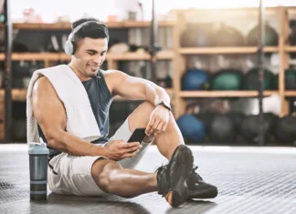 Najboljše mobilne aplikacije za ljubitelje fitnesa Muscular trainer taking a break from exercise. Young bodybuilder listening to music in headphones. Happy athlete using his smartphone in the gym. Strong, fit man relaxing after a workout