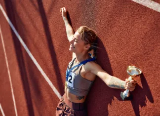Pri kateri starosti so olimpijski atleti na vrhuncu? Winner woman in sportswear with number with gold medal for first place and cup lying at stadium celebrating after competition. Sports and victory concept.