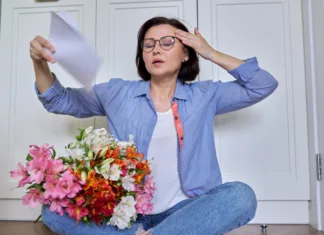 Prihaja zdravilo za vročico med menopavzo Hot flashes in a woman of mature age, symptoms of menopause, a female sitting at home on the floor blowing her face with papers