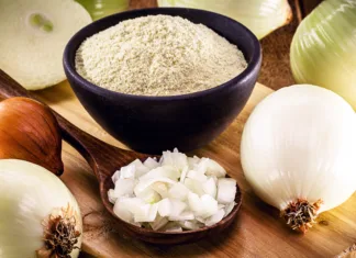 Vodič po začimbah: čebula v prahu wooden spoon with sliced onion and crushed into pieces to be used as seasoning, with powdered onion in the background, rustic kitchen