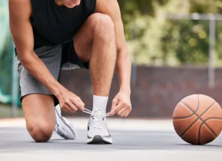 Kako izbrati prave košarkarske superge? Basketball, shoes and hands by man on the ground for lace before exercise, training and cardio at a basketball court. Fitness, sneakers and basketball player getting ready for workout, sport and game