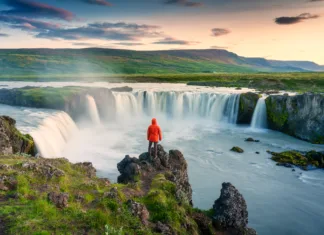 Slovenci na Islandiji: Potopis in stvari, ki jih moraš vedeti o potovanju po Islandiji Majestic landscape of Godafoss waterfall flowing with colorful sunset sky and male tourist standing at the cliff on Skjalfandafljot river in summer at Northern Iceland