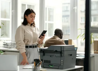 Kdaj zamenjati tiskalnik? Se da težavam izogniti? Young serious businesswoman looking at smartphone screen in office while standing by workplace with xerox machine and texting