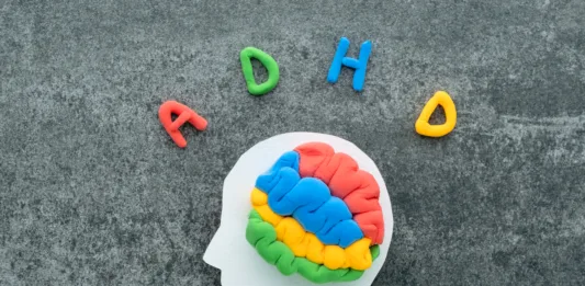 A brain model crafted from colorful materials rests on a white head silhouette, with the letters ADHD on a concrete background.