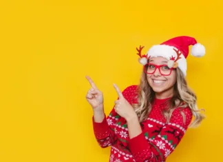 Božiček za en dan v letu 2024: Vse, kar moraš vedeti A young smiling blonde woman in a red Christmas sweater, glasses with deer antlers and a red Santa's hat points at copy space for text or design with her fingers isolated on a color yellow background