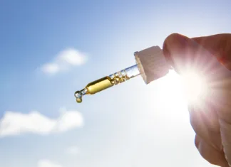 Vse o vitaminu D: Zakaj ga je predvsem v teh časih smiselno dodajati v prehrano? Hand holding dropper pipette with nice golden liquid D-vitamin against sun and blue sky on sunny day. Vitamin D keeps you healthy while lack of sun in winter, cure concept.